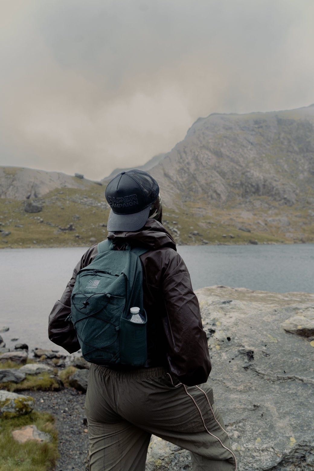 Model in a black Paper Campaign cap looking out across a mountain lake in the rain