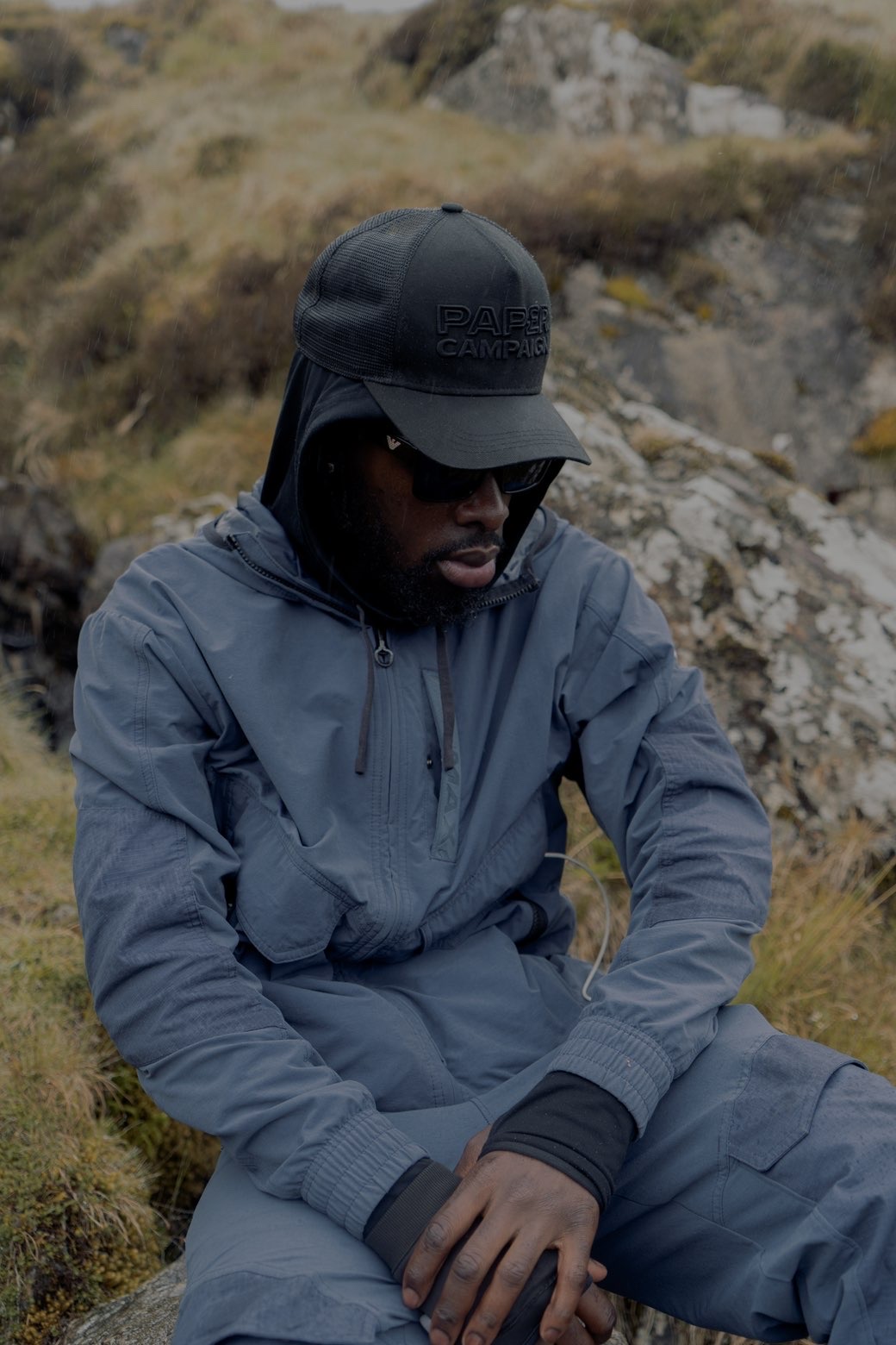 Model seated on a wet hillside in a blue-grey tracksuit wearing a dark Paper Campaign trucker cap